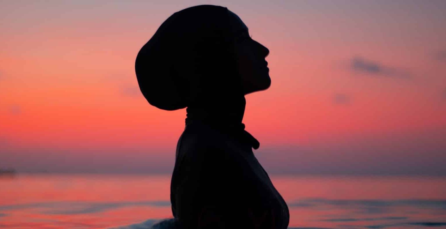the silhouette of a woman in a headscarf in chest-height water in front of a sunset
