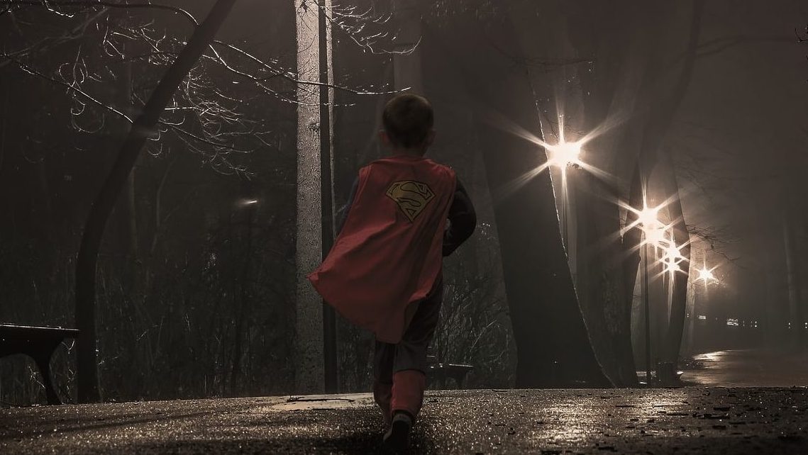 young child in a superman costume, photographed from the back, walking down a dark street with limited street light