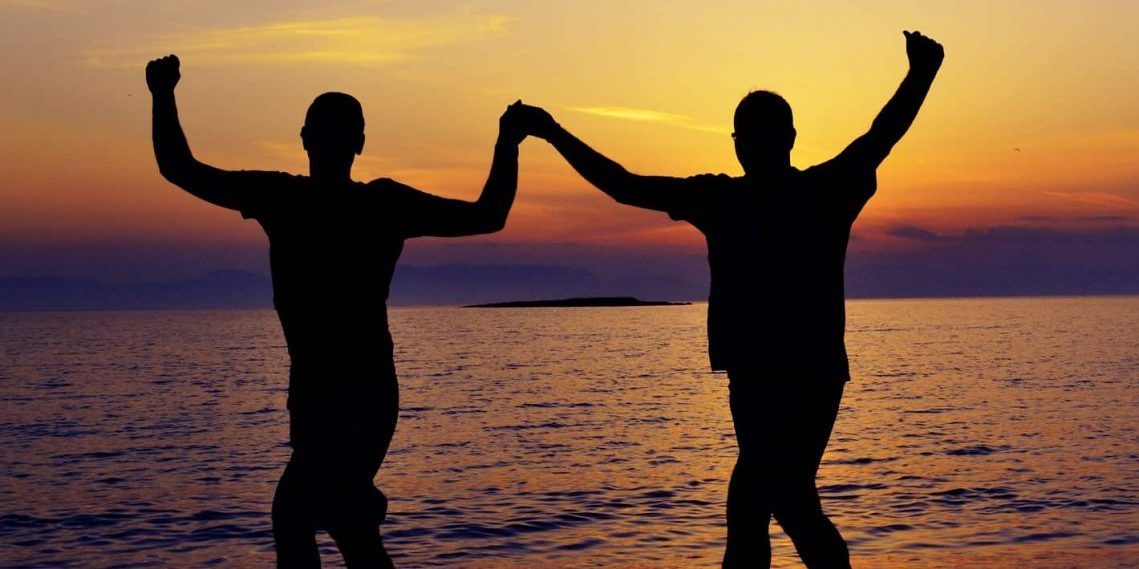two people on a beach at sunset with their hands in the air in celebration, holding one of each others hands